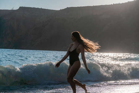 Selective focus. Happy carefree sensual woman with long hair in black swimwear posing at sunset beach. Silhouette of young beautiful playful positive woman outdoor. Summer vacation and trip conceptの写真素材