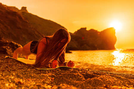 Young woman with long hair in black swimsuit and boho style braclets practicing outdoors on yoga mat by the sea on a sunset. Womens yoga fitness routine. Healthy lifestyle, harmony and meditationの写真素材