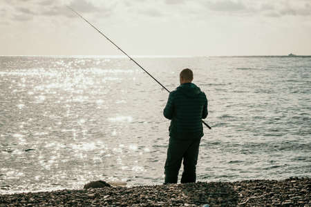 Man hobby fishing on sea tightens a fishing line reel of fish. Calm surface sea. Close-up of a fisherman hands twist reel with fishing line on a rod. Fishing in the blue sea outdoors.の写真素材