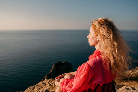 Close up shot of beautiful young caucasian woman with curly blond hair and freckles looking at camera and smiling. Cute woman portrait in a pink long dress posing on a volcanic rock high above the seaの写真素材