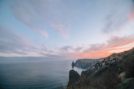 A red burning sunset over the sea with rocky volcanic cliff. Abstract nature summer or spring ocean sea background. Small waves on golden warm water surface with bokeh lights from sun.の写真素材