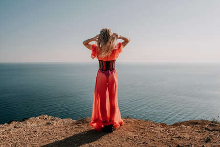 Close up shot of beautiful young caucasian woman with curly blond hair and freckles looking at camera and smiling. Cute woman portrait in a pink long dress posing on a volcanic rock high above the seaの写真素材