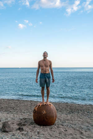 Man doing yoga exercise outdoors standing on old rusty floating marine mine on the beach with rocky shore and sea background. Healthy lifestyle, pollution, nature protection, war and peace concept.の写真素材