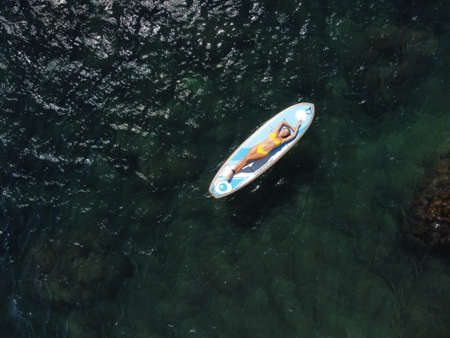 Top view on blonde woman in yellow bikini relaxing on the sup board. Young attractive woman in the sea on the Stand Up Paddle Board SUP. The concept of active and healthy life in harmony with nature.の写真素材