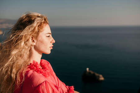 Close up shot of beautiful young caucasian woman with curly blond hair and freckles looking at camera and smiling. Cute woman portrait in a pink long dress posing on a volcanic rock high above the seaの写真素材