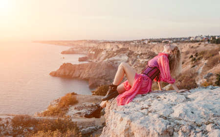 Beautiful young caucasian woman with curly blond hair and freckles. Cute redhead woman portrait in a pink long dress posing on a volcanic rock high above the sea during sunset.の写真素材