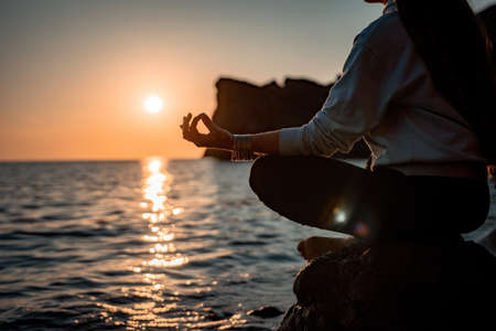Young woman with long hair in sportswear and boho style braclets practicing outdoors on yoga mat by the sea on a sunset. Womens yoga fitness routine. Healthy lifestyle, harmony and meditationの写真素材