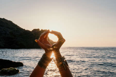 Middle aged well looking woman in white swimsuit and boho style braclets practicing outdoors on yoga mat by the sea on a sunset. Womens yoga fitness routine. Healthy lifestyle, harmony and meditationの写真素材