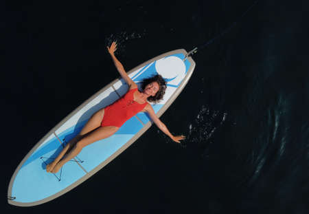 Aerial drone view on well looking middle aged woman with black hair in red swimsuit, swimming on sup in calm azure sea. Summer holiday vacation and travel concept.の写真素材