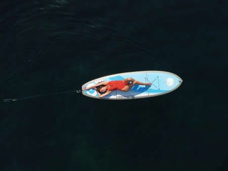 Aerial drone view on well looking middle aged woman with black hair in red swimsuit, swimming on sup...