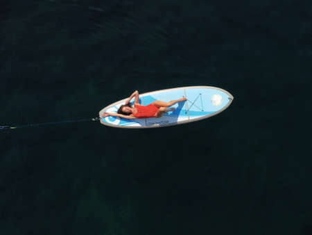 Aerial drone view on well looking middle aged woman with black hair in red swimsuit, swimming on sup in calm azure sea. Summer holiday vacation and travel concept.の写真素材