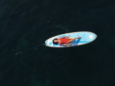 Aerial drone view on well looking middle aged woman with black hair in red swimsuit, swimming on sup...