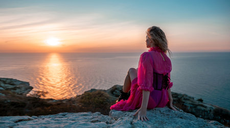 Beautiful young caucasian woman with curly blond hair and freckles. Cute redhead woman portrait in a pink long dress posing on a volcanic rock high above the sea during sunset.の写真素材