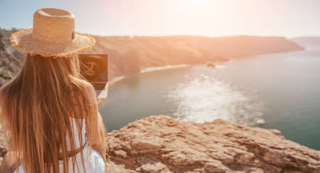 Digital nomad, woman in the hat, a business woman with a laptop sits on the rocks by the sea during sunset, makes a business transaction online from a distance. Freelance, remote work on vacation.の写真素材