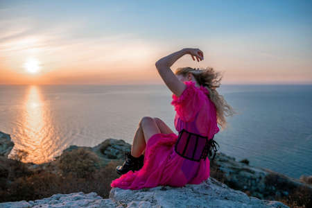 Beautiful young caucasian woman with curly blond hair and freckles. Cute redhead woman portrait in a pink long dress posing on a volcanic rock high above the sea during sunset.の写真素材