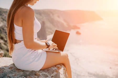 Digital nomad, woman in the hat, a business woman with a laptop sits on the rocks by the sea during sunset, makes a business transaction online from a distance. Freelance, remote work on vacation.の写真素材