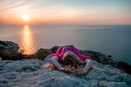 Beautiful young caucasian woman with curly blond hair and freckles. Cute redhead woman portrait in a pink long dress posing on a volcanic rock high above the sea during sunset.の写真素材