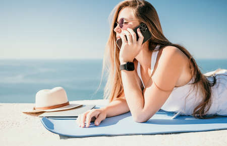 Digital nomad, woman in the hat, a business woman with a laptop sits on the rocks by the sea during sunset, makes a business transaction online from a distance. Freelance, remote work on vacation.の写真素材
