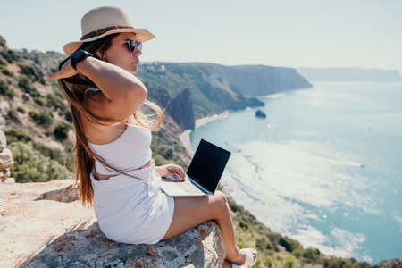 Digital nomad, woman in the hat, a business woman with a laptop sits on the rocks by the sea during sunset, makes a business transaction online from a distance. Freelance, remote work on vacation.の写真素材