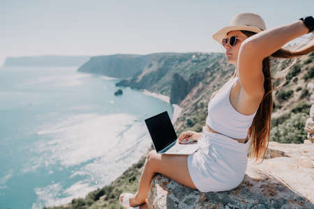 Digital nomad, woman in the hat, a business woman with a laptop sits on the rocks by the sea during...