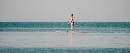 Sea woman sup. Silhouette of happy middle aged woman in rainbow bikini, surfing on SUP board, confident paddling through water surface. Idyllic sunset. Active lifestyle at sea or river.の写真素材