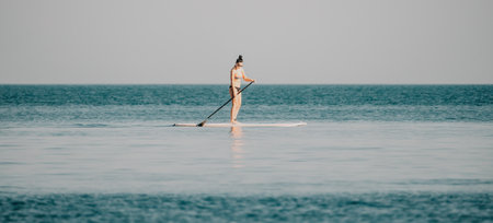 Sea woman sup. Silhouette of happy middle aged woman in rainbow bikini, surfing on SUP board, confident paddling through water surface. Idyllic sunset. Active lifestyle at sea or river.の写真素材