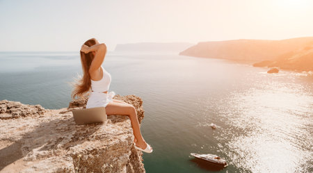 Digital nomad, woman in the hat, a business woman with a laptop sits on the rocks by the sea during sunset, makes a business transaction online from a distance. Freelance, remote work on vacation.の写真素材