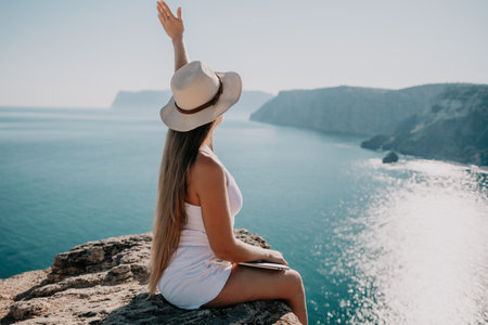 Digital nomad, woman in the hat, a business woman with a laptop sits on the rocks by the sea during sunset, makes a business transaction online from a distance. Freelance, remote work on vacation.の写真素材