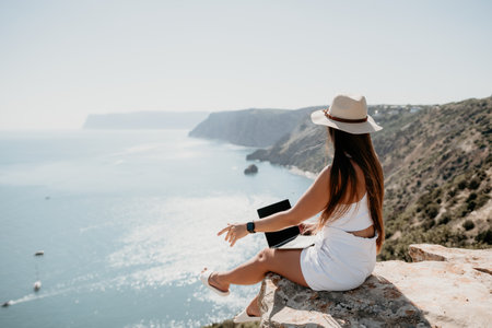 Digital nomad, woman in the hat, a business woman with a laptop sits on the rocks by the sea during sunset, makes a business transaction online from a distance. Freelance, remote work on vacation.の写真素材