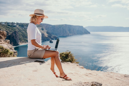 Digital nomad, Business woman working on laptop by the sea. Pretty lady typing on computer by the sea at sunset, makes a business transaction online from a distance. Freelance, remote work on vacationの写真素材