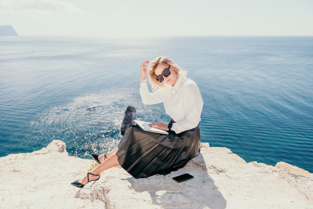 Digital nomad, Business woman working on laptop by the sea. Pretty lady typing on computer by the sea at sunset, makes a business transaction online from a distance. Freelance, remote work on vacationの写真素材