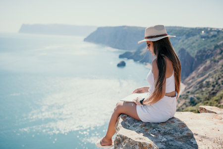 Digital nomad, woman in the hat, a business woman with a laptop sits on the rocks by the sea during sunset, makes a business transaction online from a distance. Freelance, remote work on vacation.の写真素材