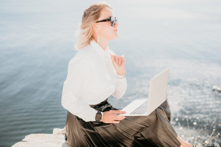 Digital nomad, Business woman working on laptop by the sea. Pretty lady typing on computer by the sea at sunset, makes a business transaction online from a distance. Freelance, remote work on vacationの写真素材