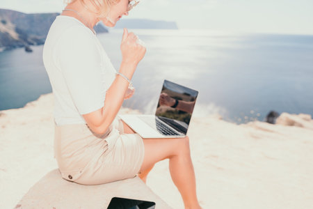 Digital nomad, Business woman working on laptop by the sea. Pretty lady typing on computer by the sea at sunset, makes a business transaction online from a distance. Freelance, remote work on vacationの写真素材