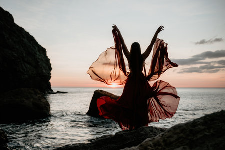 Young sensual woman in a long red dress posing on a beach near the sea on background of volcanic rocks, like in Iceland. Dreams holidays and weekend vacation in summer time.の写真素材
