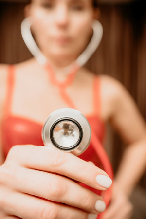 Selective focus. Young woman in red corset and black leather pants with a phonendoscope. Sexy brunette nurse in red and black costume with a stethoscope in her hands. Role doctor play. Entertainment.の写真素材