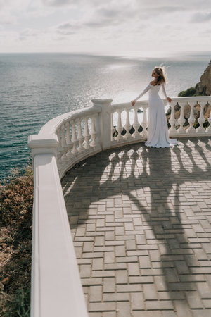 Romantic bride, blond girl in white wedding dress with open shoulders posing on open terrace with backdrop of the sea and rocks. Stylish young woman standing on terrace and looking on ocean at sunsetの写真素材