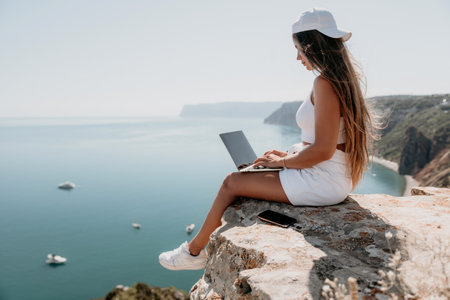 Digital nomad, woman in the hat, a business woman with a laptop sits on the rocks by the sea during sunset, makes a business transaction online from a distance. Freelance, remote work on vacation.の写真素材