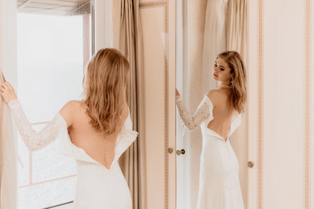 Morning preparation of the bride. Close-up of a bridesmaid, fastening a lot of buttons on the brides wedding dress. The bride in a white wedding dress with lace standing in the room. Selective focusの写真素材