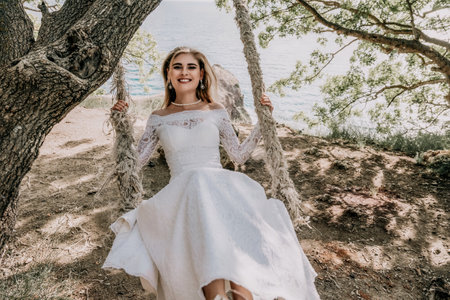 Young European beautiful girl in white bridal marriage dress posing on swing in forest trees on the ocean sea beach background. Exotic wedding photography ideas.の写真素材