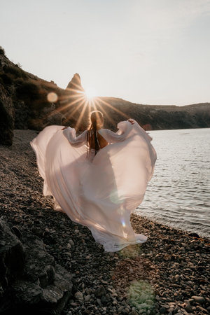 Mysterious young woman with braids in long white dress alone on the beach. Sunset over the sea with rocky volcanic cliff. Abstract nature summer ocean sea background.の写真素材