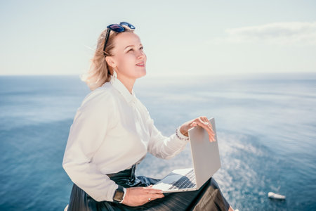 Digital nomad, Business woman working on laptop by the sea. Pretty lady typing on computer by the sea at sunset, makes a business transaction online from a distance. Freelance, remote work on vacationの写真素材