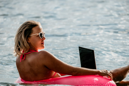 Woman works on laptop in sea. Freelancer, young blond woman in sunglases floating on an inflatable big pink donut with a laptop in the sea at sunset. Freelance, travel and holidays concept.の写真素材