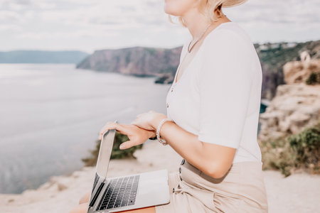 Digital nomad, Business woman working on laptop by the sea. Pretty lady typing on computer by the sea at sunset, makes a business transaction online from a distance. Freelance, remote work on vacationの写真素材