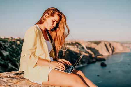 Digital nomad, Business woman working on laptop by the sea. Pretty lady typing on computer by the sea at sunset, makes a business transaction online from a distance. Freelance, remote work on vacationの写真素材