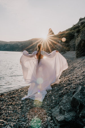Mysterious young woman with braids in long white dress alone on the beach. Sunset over the sea with rocky volcanic cliff. Abstract nature summer ocean sea background.の写真素材