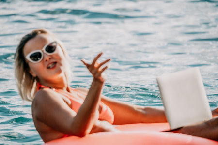 Woman works on laptop in sea. Freelancer, young blond woman in sunglases floating on an inflatable big pink donut with a laptop in the sea at sunset. Freelance, travel and holidays concept.の写真素材