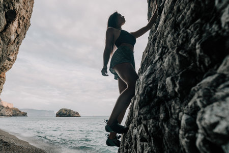 Sports Woman Climbing The Rock. Young woman With slim fit body climbing in volcanic basalt cave with beautiful sea view. The athlete girl trains in nature. Woman overcomes difficult climbing route.の写真素材