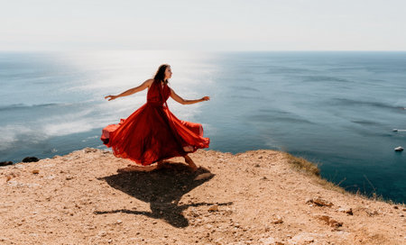 Woman in red dress on sea. Side view a Young beautiful sensual woman in a red long dress posing on a rock high above the sea on sunset. Girl on the nature on blue sky background. Fashion photo.の写真素材