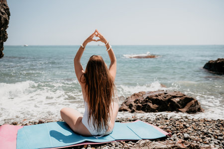 Woman makes heart with hands on beach. Young woman with long hair in white swimsuit and boho style braclets practicing by sea ocean. Concept of longing daydreaming love peace contemplation vacationsの写真素材